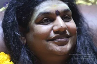 Close-up photograph of a smiling person with long dark wavy hair, a dark sandalwood or kumkum mark on the forehead, and a yellow marigold garland at the lower left edge. The background is blurred and warm-toned. Only a face and hair are visible, so the face and shoulders are the main subject.