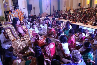 A large indoor hall with cream walls and warm wall lights holds a dense crowd of seated devotees, many women in colourful saris and salwar, some in white, pink, red and teal. In the foreground a man with long dark hair and a maroon-and-orange shawl sits on an ornate gold throne-like seat, facing a raised altar decorated with orange marigold garlands and a deity murti in crowns and jewelled attire. Devotees hold brass plates and small lamps near the front, and a long white-draped table with flower and offering plates runs along the right side. The scene suggests a puja or satsang with people seated on the floor in rows.