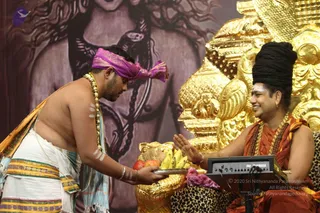 A photograph of a ceremonial offering on a stage in front of a dark painted backdrop of a goddess figure with flowing hair. A bare-chested man in white dhoti with a striped gold-and-green border and a light blue shawl, wearing a purple headband and a gold-beaded mala, stoops forward holding a silver tray of fruit, including bananas, red apples and a coconut. Seated on a golden ornate throne with leopard-print cushions is a man in saffron-orange robes with a long dark swept-back hair crown, rudraksha malas and gold necklaces, smiling and gesturing with his raised hand. A Yamaha keyboard sits beside the throne and a pink crystal rests on the cushion. The seated figure matches the Swamiji (probable) cues of long crown-style hair, saffron clothing and rudraksha malas.