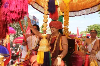 A procession scene outdoors in daylight. A person in a red-orange turban and saffron-red robe, with a rudraksha-bead and gold necklace and gold armbands, sits under a golden canopy on a decorated palanquin, smiling and raising one hand. Gold-and-green poles with long purple, red, yellow and green tassels frame the canopy, and marigold garlands hang from it. Behind, an attendant in a white dhoti and gold necklace holds a red pole, and another man in white with glasses and beads sits at the right. Crowds line the street, and a pink umbrella and red flags are visible. Several people are in view, including partially visible onlookers.