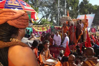Outdoor procession at a Kumbh Mela setting, with saffron-clad sadhus and devotees thronging a street. In the foreground a figure wears an orange turban and a woven orange shawl, seen from behind. A young drummer in orange robes with rudraksha malas and gold armlets plays a drum. Behind him, a decorated palanquin carries a seated figure in orange robes with a tall crown-like headdress beneath an ornate canopy, surrounded by marigold garlands, while attendants in white and saffron stand around it. Red flags, a yellow banner reading PRIMA and a white banner with Devanagari script line the route. Crowds fill the background under trees.