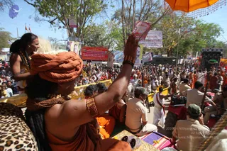 Photograph of a crowded outdoor procession during a Kumbh Mela shahi snan event. In the foreground a sadhu with an orange turban and bare upper arm raises his right hand, wearing gold-toned bangles and a rudraksha-style beaded necklace, while a woman with dark hair and gold jewellery stands beside him on a decorated vehicle with a leopard-print cloth. Behind them a dense crowd of saffron-robed sadhus, devotees and uniformed police fills the street beneath an orange canopy. Banners with Hindi text and a large portrait hang on poles and shops, and a stage with speakers stands at right.
