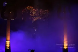A dim stage lit in violet and blue fog, with two large dark trishul (trident) forms standing on either side in front of a backdrop. Strings of warm orange fairy lights hang in a garland-like cluster above. A figure stands centrally in the smoke, mostly in shadow, with a bright face partly visible. A camera is visible at the bottom edge of the frame. The scene resembles a theatrical darshan staging with dramatic smoke and lighting.
