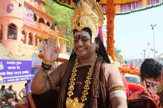 A smiling man rides in a procession under a marigold-decorated canopy, raising his right hand in a blessing wave with eyes closed. He wears an ornate gold crown with red and green jewel accents, a red bindi on his forehead, a brown-red cloth draped over his bare shoulders, heavy gold necklaces, gold armbands and bangles, and rudraksha-style beaded strands. Behind him are orange marigold garlands, a pink and orange fabric canopy, and a pink-orange building with arched windows. Police personnel and a police assistance booth with purple signage are visible at the left, with a crowd and roadside barriers in the background. The page's a person flag is no, so no identification is made from appearance.