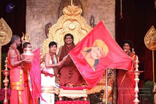 A seated man in red-saffron robes and a rudraksha mala sits on an ornate gold throne with carved peetha-style backrest, centred on a stage with dark red curtains. On his right, a man in white dhoti with a red-and-gold shawl holds a red flag printed with a portrait labelled in no legible text, its crest-like print showing a smiling figure in a yellow turban. Beside him a young man in saffron dhoti holds a silver kalasha pot, and a second attendant in saffron stands at right holding a ceremonial brass lamp stand and a clay ceremonial item. Gold ceremonial umbrella-finials and a trident stand near the throne.