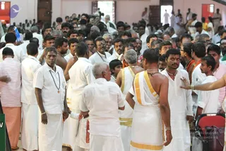 A dense crowd fills an indoor hall with pale pink walls and red doorframes. Most men wear white dhotis and white shirts; some wear white shirts with yellow-bordered dhotis, and a few wear saffron or orange cloth. Several men wear rudraksha malas and beaded necklaces, and one has a red tilak on his forehead. Older men with grey hair stand among younger men, and a child in a white shirt is visible near the centre. Men and women stand packed together, with a red baby stroller at the lower right and a green sign at the left edge.
