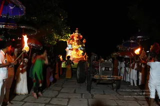 A night procession on a stone-paved path. At the centre, a brightly painted Ganesha murti with a red crown and blue-and-gold ornaments sits on a decorated trailer pulled by a red tractor. Torch-bearers hold flaming torches near the front on the left, and a man in a white shirt and a woman in a white sari with gold border walk alongside. Ornate umbrellas with mirrored, sequinned edges in blue and red line the path. Devotees in saffron and white clothes follow the trailer, and a man in white stands at the right holding a camera. The scene is lit by the flames and the glowing murti.