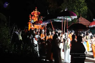 Night procession with a large illuminated Ganesha murti on a float at upper left, painted in red, orange and gold with a crown and elephant trunk. Beneath it, devotees in orange and saffron robes and white dhotis walk among red and purple decorated umbrellas on long poles. A woman in white sari stands at center, and a bare-chested man in orange garment with rudraksha beads appears at right, holding a ceremonial fan. Trees and a tiled-roof shrine line the dark background.