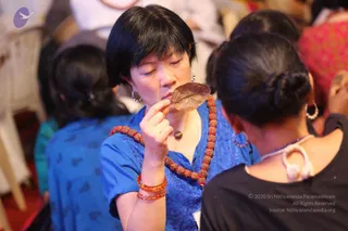 A woman in a blue short-sleeved top with a red rudraksha mala is seated in a crowd, eyes closed, holding a dried brown leaf to her lips with her right hand. She wears orange and red bangles and drop earrings. Behind her and beside her, other seated attendees in blue, black and red clothing are visible, with white plastic chairs in the background.
