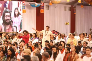 A large indoor gathering of devotees seated on white floor covers under hanging colourful paper lanterns and a red-and-white draped stage backdrop. A woman in the centre wears a cream salwar with a red-brown rudraksha mala and arms outstretched, while a woman in a red top beside her raises her hands. Many attendees wear white, orange and saffron clothes and malas. A large screen on the left shows a bearded man in saffron with a rudraksha mala, and a digital clock reads 11:13.