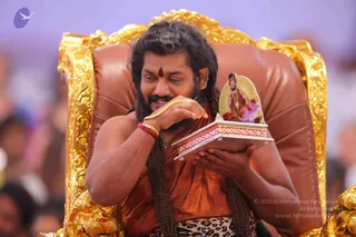 A man with long dark hair and a beard sits on an ornate gold-carved throne with brown leather upholstery. He wears an orange-saffron garment, a red tilak on his forehead, a red and gold wristband and a gold bangle, and a leopard-print cloth at his waist. He smiles while holding a small wooden box with red and gold trim, on its lid a printed picture of a seated figure. A blurred crowd fills the background. The image is a photograph.
