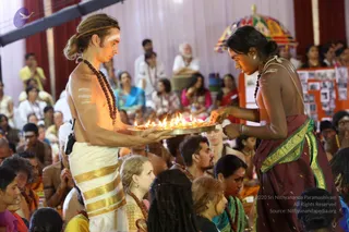 Photograph of a ceremonial aarti at an indoor event. In the foreground a shirtless man with long hair tied back and white ash markings on his chest and arms, wearing a white dhoti with gold border and a rudraksha mala, holds a brass tray of lit oil lamps. Opposite him a younger person in a maroon sari with green and gold border and a rudraksha mala reaches toward the flames. Seated devotees fill the hall behind them, and a decorative umbrella and a wall of framed photographs appear in the background.
