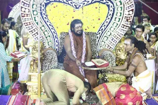 A photograph of a ceremonial event in a decorated hall. A man seated on a golden throne with a leopard-print cushion wears a saffron-orange robe and a pink and white flower garland, smiling while holding a red-covered book on a golden tray. Behind the throne a large heart-shaped backdrop of yellow flowers and ornate patterned trim frames him. To the right, a bare-chested man with long dreadlocks and white dhoti pulled from a tray a red-covered book and offers it. In the lower foreground, a man in tan clothing bows deeply with his head near the floor, and a woman in a red and gold sari kneels nearby. Standing attendants in white, gold and teal clothing surround the platform, and a tall golden ritual staff stands at left. Seated and standing devotees fill the background.