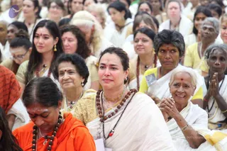 A large seated audience at a Guru Purnima gathering, mostly women in white and cream sarees and kurtas with yellow and gold accents. In the foreground, a woman in a cream and gold saree with a long rudraksha mala and a gold pendant looks tearful, with a tilak on her forehead. Beside her, an older woman in white holds her hands in anjali and smiles, and another woman in yellow wears a rudraksha mala. At lower left, a shaven-headed woman in a saffron-orange robe with a rudraksha mala sits with eyes lowered. Behind them, younger and older devotees sit close together in the hall. The scene reflects satsang and discourse seating.