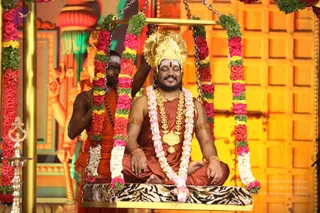 A photograph of a seated man on a gold swing decorated with red roses, yellow and white flowers and green foliage, hung from chains. He wears a large gold crown, a gold chest ornament and multiple gold necklaces, pink-and-white floral garland, gold armbands and a saffron-orange robe, with a red tilak on his forehead and eyes closed in meditation. Behind him a man in saffron-orange robes and rudraksha beads holds one swing chain. A brass oil lamp stands at left, with gold and orange backdrop panels and a tiger-striped cushion on the swing seat.
