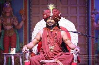 A seated man in maroon-red robes and a matching red turban with a yellow feather and jewelled ornament sits on an ornate white-and-red throne, eyes closed and smiling, with a microphone on a stand before him. Rudraksha-style beaded malas and a large gold pendant hang at his chest, with gold bangles on his upper arms. Silver lion-head armrests flank the throne. To the left stands a painted deity murti with multiple arms and a red-and-green dhoti, and a small framed photo sits on a silver side table. The background is a carved wooden panel. Swamiji (probable): long dark hair, turban, red robes and rudraksha malas indicate the presenter at a satsang on a throne.