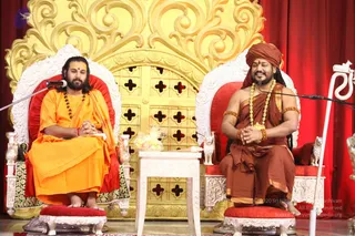 Two men sit on ornate white-and-red thrones before a gold-patterned backdrop with red curtains. The man on the left wears a bright orange robe, a rudraksha mala, and has long dark hair and a beard; his hands are folded and he is seated behind a microphone. The man on the right, seated on the right throne, wears a rust-brown drape with gold trim, a gold-beaded ceremonial necklace, gold armlets, and a brown turban; he smiles with hands clasped in front of a microphone. A small white table with a gift basket sits between them. Swamiji (probable) is the man on the right, identified by the turban, gold necklace, armlets and ceremonial brown drape alongside the throne seating and microphone; the other seated man is a guest.