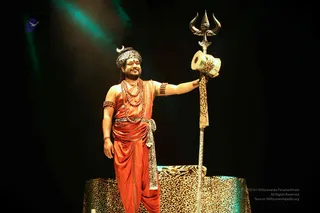 A man stands on a leopard-print raised platform on a dark stage, lit by a teal spotlight. He wears an orange-red draped garment, a leopard-print headwrap with a small crescent ornament, a multi-strand rudraksha mala, and rudraksha bracelets. He holds a tall silver trident with a leopard-print tassel and a small drum at its base. a person: the figure wears a leopard-print headwrap, rudraksha malas and ornate ceremonial dress, consistent with the page's Inner Awakening event context. A small blue bird logo appears at the upper left of the frame.