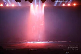 A photograph of a dimly lit stage with red and pink light beams falling from overhead lamps onto a haze-filled central area. A faint silhouette of a standing figure holding a tall staff or trident is visible in the centre, partly obscured by smoke. Large dark panels frame the stage, and the floor is lit with a reddish glow. The figure is too indistinct to identify, so no identity is claimed.
