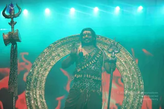 A man stands on stage in front of a large ornate gold circular arch, against a teal-lit backdrop with red flame-like shapes. He wears a leopard-print garment with a belt of brass bells, a rudraksha-style necklace, and long hair swept back, and holds a silver trident in his right hand while raising his left palm. Beside him a tall ornate staff topped with a trident and a small drum is hung with a leopard-print cloth. Swamiji (probable): long swept-back hair and a leopard-print garment with a bell belt and a rudraksha-style necklace are visible, and he is performing as a central figure in the stage presentation. Stage lights are visible overhead.
