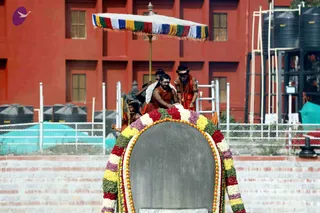 A photograph of a ceremonial procession on a brick-paved platform in front of a red multi-storey building with water tanks. A large dark stone structure shaped like an arched tomb-like shrine is wrapped in a garland of marigolds, roses and green leaves in red, yellow, white and purple. A striped canopy umbrella with red, green, blue and white fringes is raised above it on a pole. Several men stand on and around the shrine: one in the centre wears an orange-brown robe and holds the shrine's edge, one wears a white shawl and a head covering, and one on the right wears an orange robe, rudraksha beads and a headband. Their gestures suggest a ritual or ceremonial offering at the shrine.