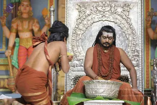 A seated man in saffron-orange robes, with long dark hair, a white vibhuti line on his forehead, and multiple rudraksha mala necklaces, sits on an ornate silver throne with a leopard-print cushion. Before him, a kneeling attendant with a bare back and an orange dhoti, wearing a dark head wrap and beaded wrist bands, offers something at a large silver bowl set on a green banana leaf. Behind them stand painted deity figures with multiple arms and gold ornaments, and a silver ceremonial staff stands at left. The scene appears to be a puja or abhishekam ritual in a temple-style hall.