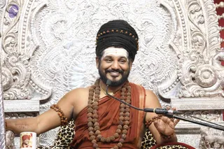 A smiling bearded man sits on an ornate silver throne with intricate repoussé floral and scroll carvings, speaking into a clip-on microphone on a stand. He wears a long black turban-style hairstyle wrapped with rudraksha beads, a white horizontal tilak on the forehead, a saffron-orange shawl over a bare upper body, multiple large rudraksha mala necklaces and a thin gold chain, and rudraksha bracelets on both arms. He gestures with his right hand. A leopard-print cushion lies beneath him. A mug with a small printed portrait sits at the lower left. Only one physical person is visible.