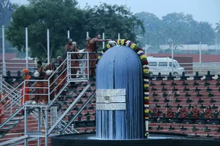 A large dark blue cylindrical Shiva lingam stands on a black circular base, decorated with a multicoloured flower garland of marigolds in yellow, orange, purple and white draped over its top. On the metal scaffolding stairs and platform to the left, several men in saffron and orange robes with rudraksha malas stand and pour liquid and handle white vessels, one man in orange garments reaching toward the lingam. Below, rows of seated monks in saffron robes sit on tiered red brick steps, some holding small items. A white van and palm trees appear in the background under a pale sky. This is a photograph of an abhishekam ritual at a lingam.