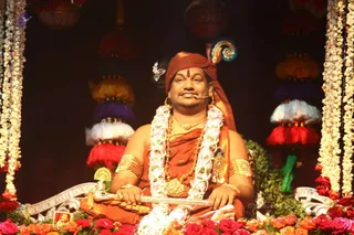 A seated man in saffron-orange robes and a rust-red turban sits on an ornate silver throne decorated with fresh flowers and greenery. He wears a white and pink flower garland, gold necklaces, rudraksha beads and gold armbands, and a small crest is pinned at his turban. He holds a silver scepter-like rod across his lap. Red and white tassel decorations and white flower strands frame the throne, with red and orange roses at the base. a person: the figure wears distinctive ornate saffron clothing, rudraksha malas and gold ceremonial jewellery in a festive setting; the page identifies the event as a Krishna Janmashtami celebration.