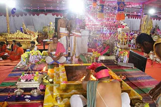 A festive temple hall during Navaratri with a yellow-and-gold homa or puja fire pit in the foreground, flames rising from a wooden platform decorated with pink flowers. A bare-chested priest in a red-pink headcloth and white dhoti bends over the fire, and a second priest in white dhoti stands at right. Two standing men in white dhotis with colourful borders stand at the centre. Seated devotees in orange and white clothes sit on striped mats at left. Behind them are tiered displays of small figurines and lamps, a golden palanquin or throne, a deity murti, and fruit, flowers and brass vessels on the floor. Coloured lights and hanging decorations fill the ceiling.