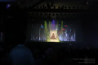 Photograph of a large darkened auditorium viewed from the back, with silhouetted audience members in the foreground, including a person in a pale top at left. On the stage screen, a projected scene shows a seated figure in orange-red robes on an ornate golden throne, flanked by tall ornamental pillars or temple-like structures with red and green lighting and green light beams. Purple and blue stage lights hang from the ceiling rig. The figure is on screen only, so the candidate is a depiction; identity is uncertain from appearance alone.