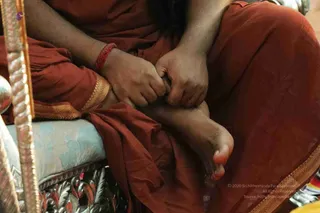 Close-up of a seated person in a rust-orange robe on an ornate silver-framed chair with a pale blue floral cushion. Their hands rest on a bare foot, and a red thread is tied around one wrist. Gold-bordered fabric trims the robe, and a golden carved armrest edges the left side. The frame is cropped at the knees, so no face is visible and identity cannot be judged from the image.