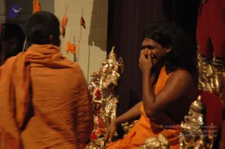 A photograph taken indoors in dim light. A seated man in saffron-orange robes with long, swept-back dark hair laughs, one hand raised to his face near his nose. He sits beside an ornate gilded metal deity figure with red fabric accents, and a silver ceremonial pot is visible at the lower right. In the foreground a second man with a shaved head, wearing an orange shawl draped over his shoulders, is seen from behind. Orange flags on thin poles stand against a pale tiled wall behind them, and red drapery frames the right edge. Swamiji (probable): the seated man wears a saffron robe, has long swept-back hair and sits in a ceremonial setting beside gilded ritual objects.