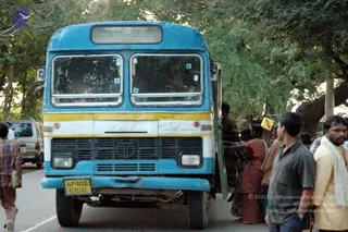 A photograph of a blue and white Tata bus parked beside a tree-lined road, with its front door open. Passengers, including women in red and maroon saris, are boarding or standing by the door. On the right a man in a green shirt and a man in a yellow shirt with a white cloth over his shoulder stand on the roadside. A silver van is parked behind the bus on the left, and a man in a checked shirt walks on the left edge. The bus has yellow stripes and a yellow number plate reading AP 107 6106.