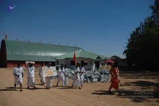 A dusty dirt road in front of a green corrugated-roof building with a red wall and a whitewashed structure beside it. A procession of about seven men in white dhotis and white shirts walks toward the camera; the front man carries a framed picture on a board, another carries a bundle on his head, and one carries a small orange flag on a pole. Striped stone blocks are piled beside the whitewashed building. On the right, a figure in an orange-saffron robe with a garland and a walking staff walks behind the group, with a clear blue sky overhead.
