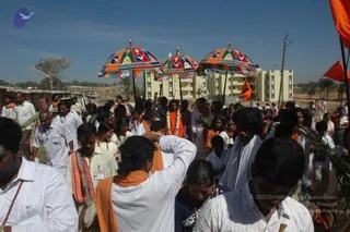 Outdoor procession on a dusty open ground with a clear blue sky. Three large ornate umbrellas with multicoloured panels, gold finials and fringed edges are held aloft over the crowd. Many people in white shirts and white dhotis, some wearing orange shawls, walk closely together. A man in the centre wears a white garment and a saffron-orange garland. Saffron flags and palm fronds are visible, and a row of low apartment blocks stands in the background. Several people carry small objects and one man on the left wears a rudraksha mala.