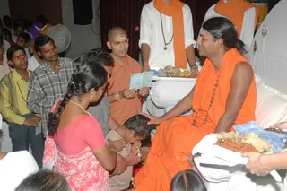A seated man in saffron-orange robes with long swept-back hair and a rudraksha mala sits on a white-draped throne, gently touching the head of a small child kneeling before him. Next to him, a shaved-headed monk in orange robes with a tilak holds up a printed receipt or card. A woman in a pink sari with a long plait and gold necklace bows with hands folded, and a man in a checked shirt stands in line. Behind them, attendants in white shirts and orange shawls stand near a table with flower offerings, fruit and a tray of kumkum-coloured prasad. Devotees fill the room, and a pedestal fan and red curtain are visible in the background. Swamiji (probable) is the seated figure receiving devotees, identified by the long swept-back hair, saffron robes and rudraksha mala.