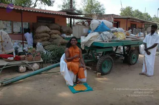 Outdoor ashram scene on a dusty ground beside a low orange-walled building with a corrugated roof. A smiling person with long hair, a red-orange forehead mark and an orange saffron-coloured robe sits on a folded teal mat with a yellow patterned design, one leg bent, beside a green-painted handcart piled high with white sacks, bags, cloth bundles and boxes. Stacked jute sacks sit behind the cart. To the left, a table holds terracotta pots and a red cushion, with white garland-decorated fabric nearby. A man in a white shirt and white dhoti stands at right, and another person in white is partly visible at left. Green pipes lie on the ground.