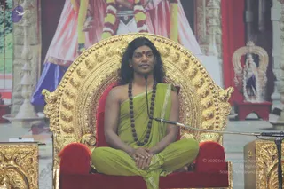 A man sits cross-legged on an ornate golden throne with red velvet cushions, hands folded in his lap, at a microphone on a stand. He wears a green saffron-toned draped garment over bare shoulders, a long dark rudraksha mala around his neck, and a small red tilak on his forehead. Behind him a large backdrop shows a figure in pink and white robes with a garland, and a smaller golden shrine with a deity murti on the right. Gold carved pillars frame the stage. Appearance-based candidate: Swamiji (probable), seated on the throne in a satsang setting, identified by long swept-back hair, rudraksha mala and ceremonial throne cues.