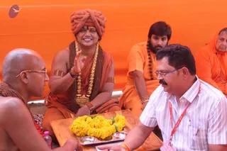 A photograph taken at an outdoor gathering in front of a bright orange backdrop. A man in an orange turban and saffron-orange wrap sits cross-legged, wearing a long gold necklace and a rudraksha mala, with gold armbands and a red bracelet; he smiles and raises one hand in blessing. Beside him, a bald man in a plain saffron wrap and rudraksha beads sits with his hands near a wooden tray holding a yellow marigold garland and small metal bowls. Opposite, a man in a white striped shirt, glasses and a red lanyard leans forward with hands extended toward the tray. Two more people in orange robes sit in the background, one with a rudraksha mala. Swamiji (probable): the seated figure in the turban shows distinctive saffron clothing, turban and ornate gold necklace cues.