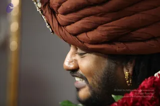 Close side-profile photograph of a smiling man wearing a large rust-brown silk turban with a small gold and green ornament at its edge. He has gold hoop earrings, a short beard and moustache, and eyes gently closed. Red flowers and green leaves. The background is a soft grey blur with a golden vertical pillar at the left.