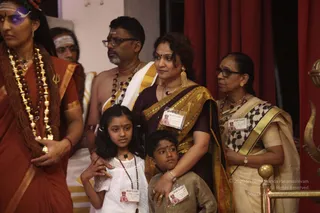 A photograph of a group standing before red curtains at an indoor event. At the far left, a woman in an orange-red sari with a gold-and-bead garland and a rudraksha-style necklace stands beside a partly visible person wearing a white forehead band. In the centre, a bare-chested man with a sacred thread, a grey-streaked beard, glasses and a vibhuti mark wears a white dhoti with gold border and rudraksha mala. Beside him, a woman in a maroon and gold sari with gold earrings and necklace holds a girl in a white dress with a beaded necklace and a boy in a beige kurta; all three wear name badges. At the right, an older woman in glasses and a cream sari with gold border and black-and-white patterned pallu stands near a brass railing with a trident finial. Several visible people wear identification badges.