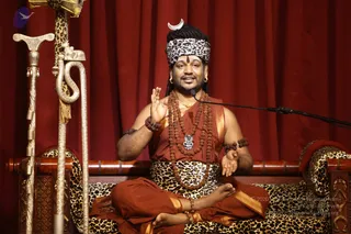 A seated man in orange-red robes sits cross-legged on a leopard-print cushioned throne in front of a deep red curtain. He wears a leopard-print headband with a silver crescent, multiple rudraksha malas, a silver pendant, and beaded bracelets, and gestures with his right hand raised while speaking into a microphone on a stand. A gold-tipped staff with a carved serpent and a ceremonial stand stand to the left. Appearance-based cues (long hair under headband, ornate mala and pendant, saffron-orange robes) suggest a probable Swamiji (probable) giving a discourse.