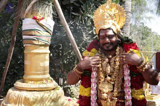A photograph of a man with eyes closed and hands pressed to his chest beside a golden lingam-shaped pillar wrapped in white and gold cloth with a thatched cover above. He wears an ornate gold crown, a thick garland of red and yellow flowers with pink strands, gold necklaces, bangles and rings, and a vibhuti mark on his forehead. Flowers are scattered at the base of the golden pedestal. Green foliage and a palm frond fill the background.