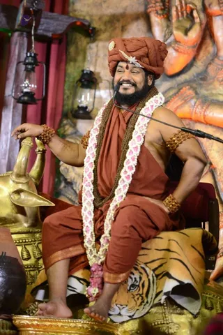 A smiling bearded man sits on a carved wooden seat draped with a tiger-striped cloth, speaking into a microphone on a stand. He wears a rust-orange turban with a silver ornament and a red tilak mark, a rust-orange robe, and a brown knotted shawl. A long garland of white and pink flowers hangs across his chest, and he wears rudraksha bracelets on his wrists and arms. His left hand rests on a golden elephant-head armrest. Behind him are a large orange carved figure and two hanging lanterns. Appearance cues are consistent with Swamiji (probable): a turban, ornate orange clothing, rudraksha malas and a garland, seated as a speaker on a dais.