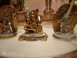 Close-up photo of small gold-toned metal murtis on a white marble surface. In the centre sits a seated Ganesha figure with an elephant head, holding objects in his hands and resting on a lotus-like base. Flanking him on both sides are winged, flame-backed figures with ornate scrollwork, one with a crown. Candle wax drips over the bases. Blurred in the background are a standing bronze figure, a brass lamp and fairy lights.