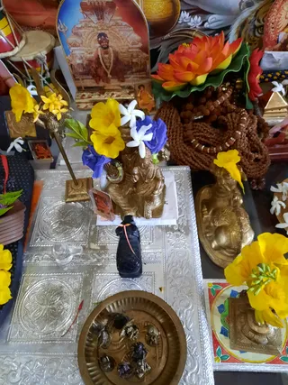 Overhead photo of a home altar with a silver-embossed cloth. At the centre sits a small brass deity murti decorated with yellow and white flowers and a blue flower, beside a black stone lingam with a red thread. A brass plate holds dark dried offerings. A framed printed portrait of a seated figure in a saffron-coloured garment and a beaded necklace stands at the top left, with a brass lotus-shaped arrangement of orange flowers and rudraksha malas at the upper right. A large brass oil lamp, drums and a brass kalasha-like object appear at the edges. No people are visible.