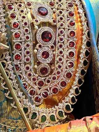 Close-up photograph of an ornate deity murti or decorated figure adorned with a heavy gold necklace set with large red stones, smaller red and white stones, pearls and green stones, layered over a teal and gold brocade garment with an orange silk cloth beneath. A metal staff or rod with a gold tip is visible at the left edge. Ceremonial jewellery dominates the frame.