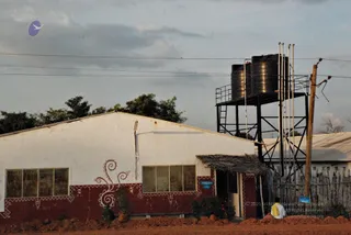 A rural building with white upper walls and a maroon lower band, decorated with white painted scrollwork and border patterns. A thatched-roof canopy sits over the doorway. Beside it stands a tall black steel water tank stand holding two dark cylindrical water tanks, with metal pipes rising alongside. A wooden fence, utility poles with power lines and a red earth path lie in front, under a cloudy evening sky. A single figure in a white garment with a yellow shawl walks along the path at the lower right edge. A small sign hangs beside the doorway and a purple bird logo appears at the upper left.