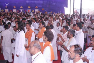 A large crowd of men in white ritual clothes stands in a tented hall with a blue canopy, many raising their arms and clapping. Several men in front wear saffron-orange shawls over white kurtas; one has long dark hair and wears white, another has curly hair with an orange shawl. A bald older man in white wears a red-beaded mala. Along the back wall, framed portraits of saintly men are decorated with orange and yellow marigold garlands. A banner at upper right is partly legible. a person is not clearly identifiable by appearance, so no candidate is named.